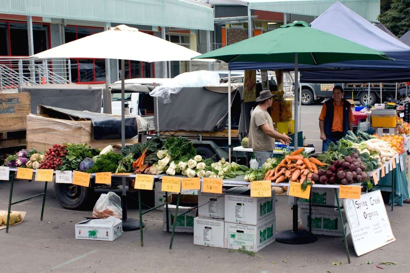 Busy outdoor farmers market with fruit and vegetable stalls under umbrellas